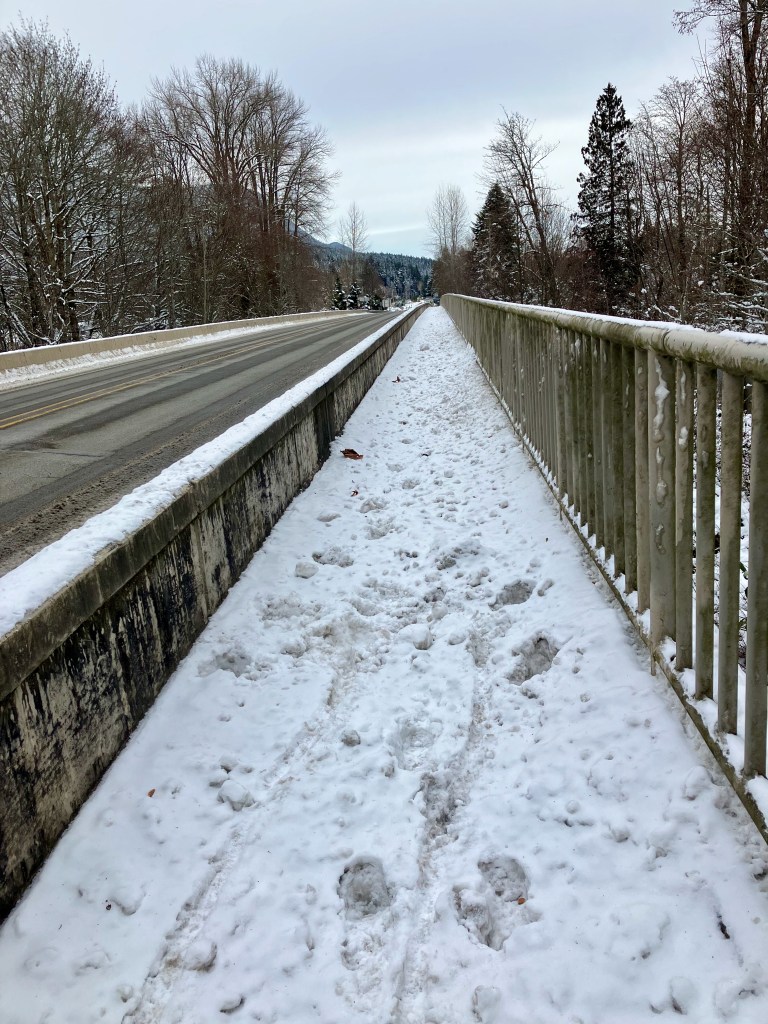 The snow-covered pedestrian walkway along US 101 crossing the Dosewallips River in Washington. There are footprints and two tire tracks. 