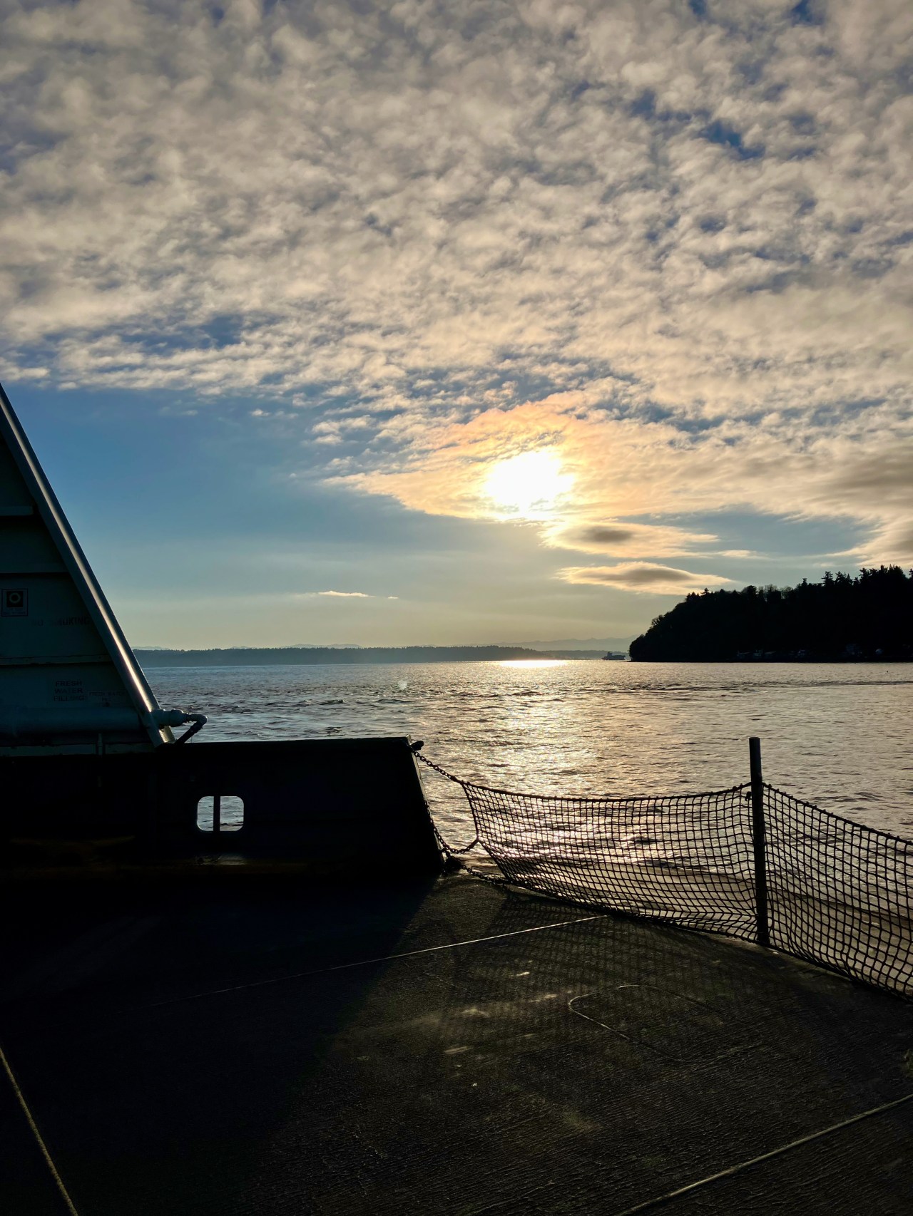 Bright morning sun radiating through a cloud in an otherwise blue sky over a Puget Sound seen from the deck of a ferry about to land at Vashon island.