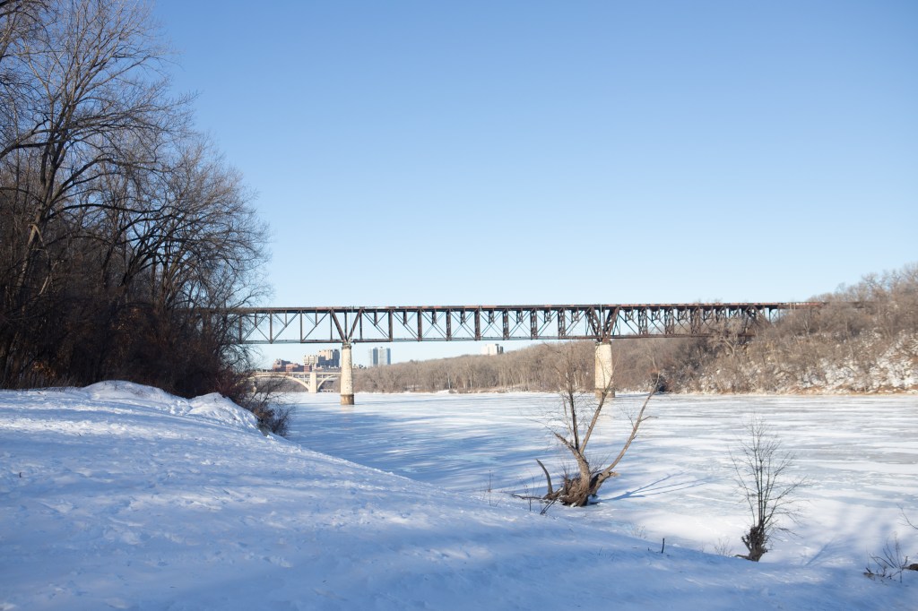 A river covered in snow with a railroad bridge in the near distance and in the far distance a few skyscrapers.